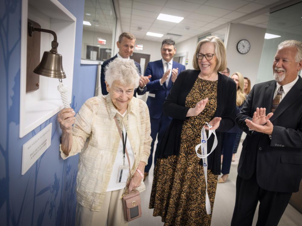 An older woman smiles as she rings a ceremonial bell mounted on the wall at the opening of the Penn State Health Cancer Center at Hampden Medical Center.