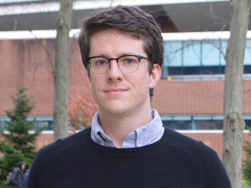 Harrison Cole, a person with short brown hair and glasses, wearing a black sweater over a collared shirt, stands outdoors in front of a brick building and trees.