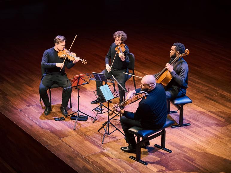 Two white and two black men sit on a stage and play stringed instruments.