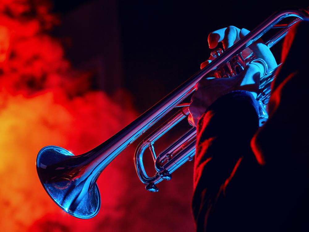 Close-up of a person playing a trumpet, illuminated by dramatic blue and red stage lighting with smoky background effects.