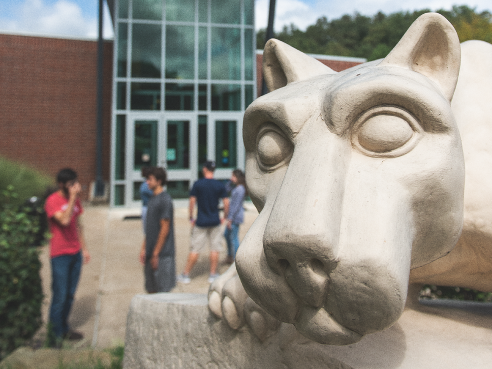 Close-up of the Nittany Lion Shrine statue at Penn State Greater Allegheny, with a group of students talking in the background outside a campus building.