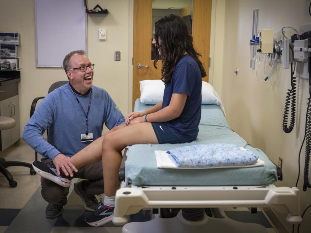 Dr. Matthew Silvis smiles as he looks up at a teen girl sitting on an exam table. He is pressing down on her ankle. He is wearing a long-sleeved shirt and a nametag with a lanyard. The girl, who is talking, has long hair and is wearing a T-shirt, shorts and sneakers.