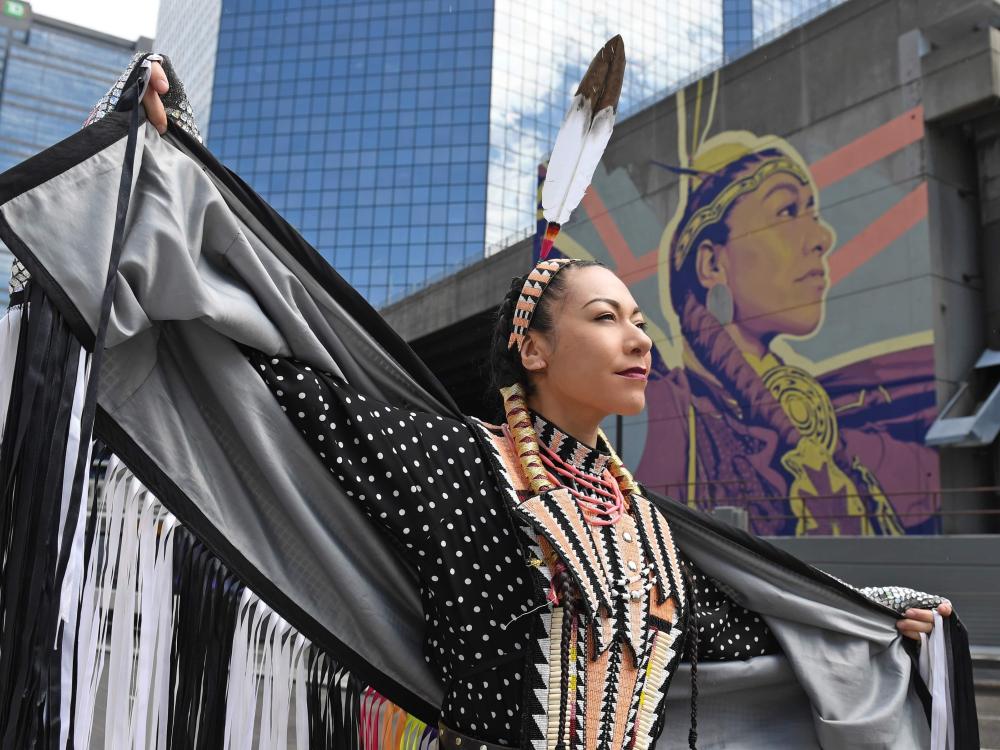 A Native American woman stands in front of a mural of an indigenous chief.
