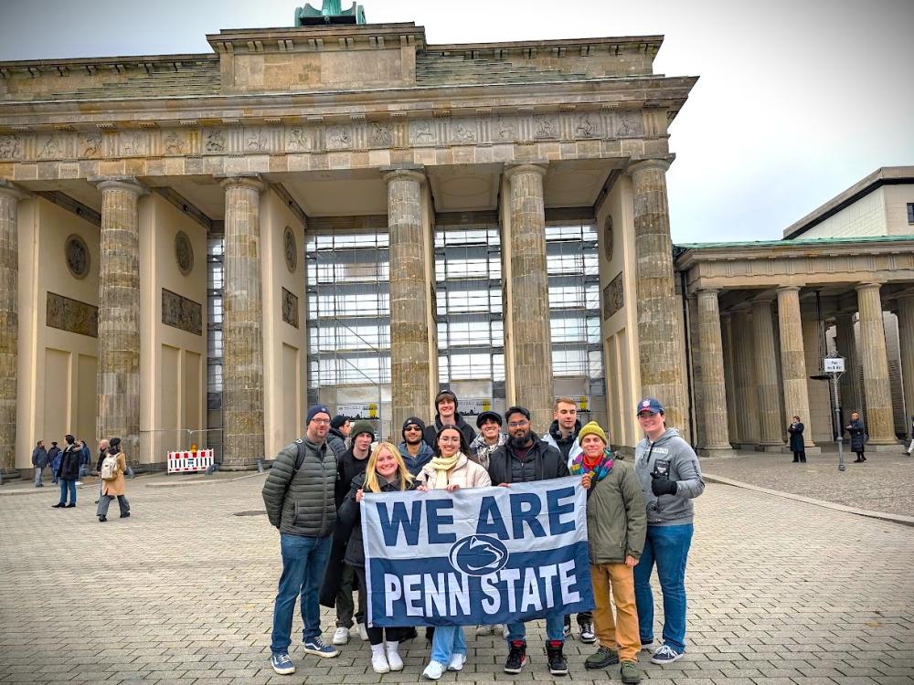 A group of students posing with a "We Are Penn State" flag in front of a building