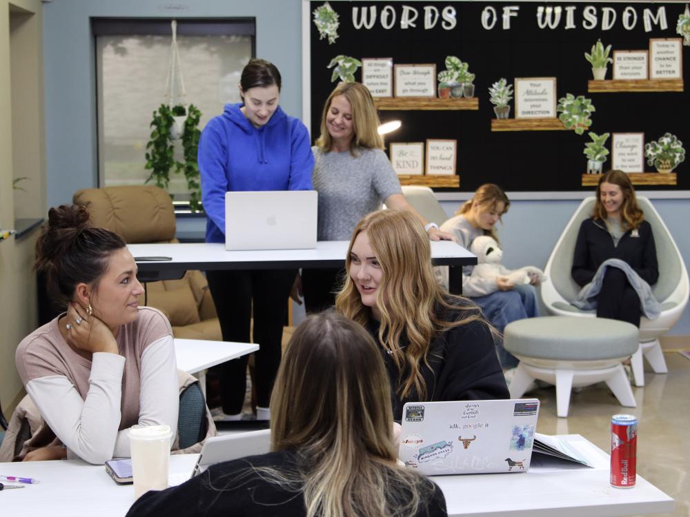 Students and faculty sitting at tables and standing at desks around a classroom with laptops and books