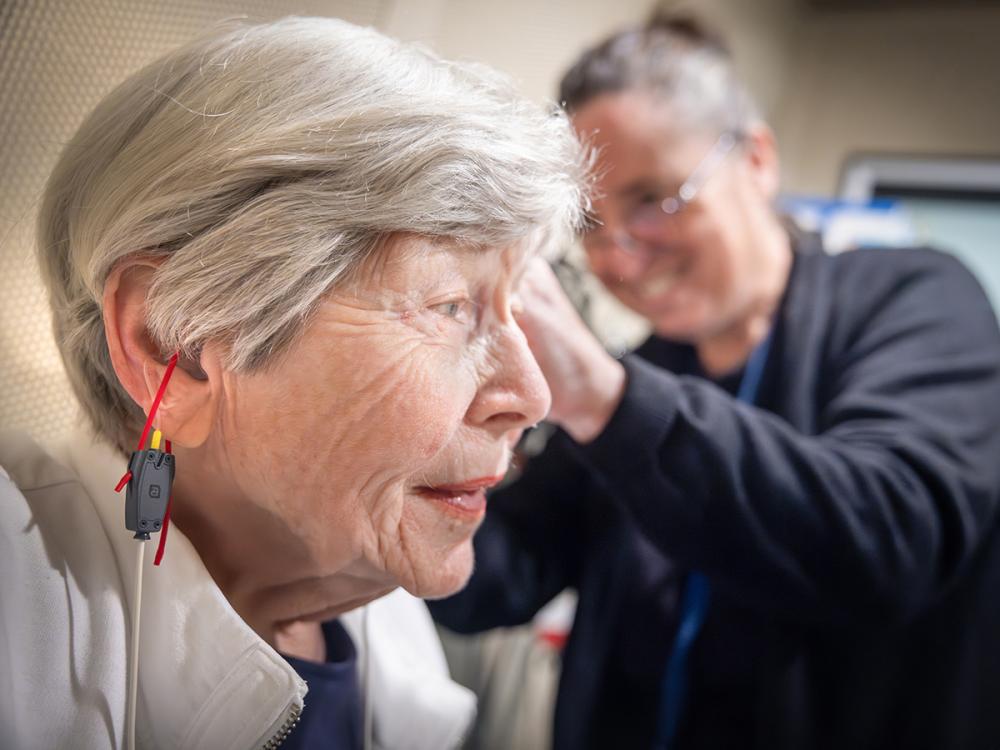 A woman is shown in profile as Dr. Jackie Price, an audiologist at Penn State Health Otolaryngology – Head and Neck Surgery, fits her with a hearing aid. A cord is hanging from the woman’s ear, and small ribbons hang from the hearing aid. Price, who is seen out of focus, is wearing scrubs, a lanyard and glasses and is smiling.