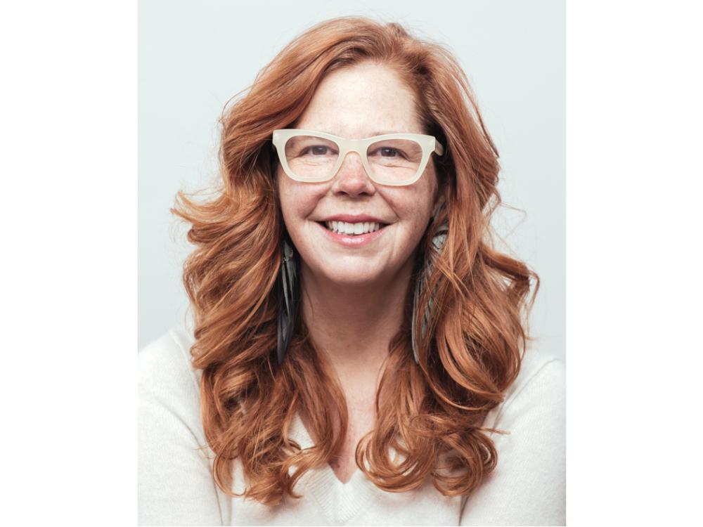 An individual with red hair and white-framed glasses against a white background.