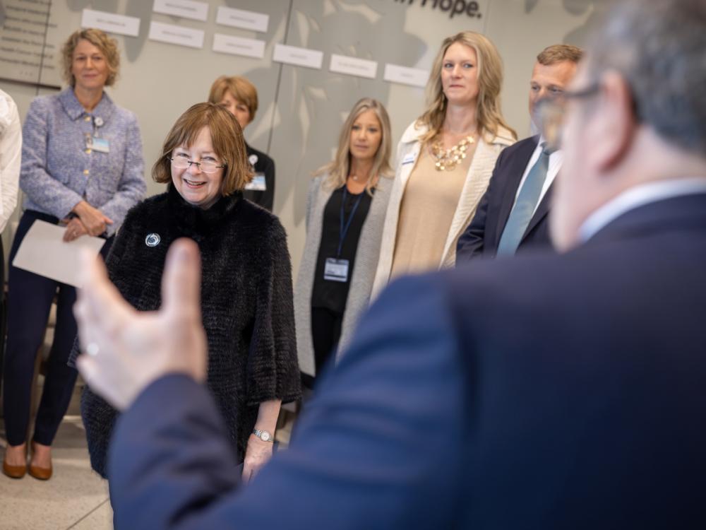 A group of people form a semi-circle around a woman who smiles as she looks at a man in a suit standing near the camera.