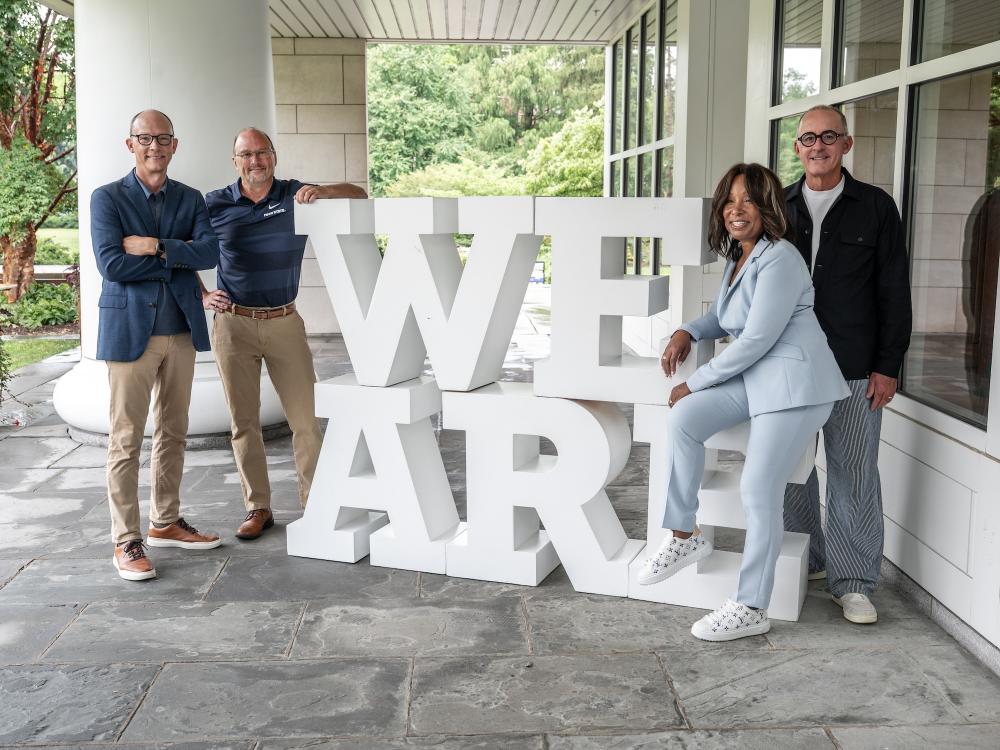 Four people gather around a "We Are" sign on the porch of the Hintz Family Alumni Center.