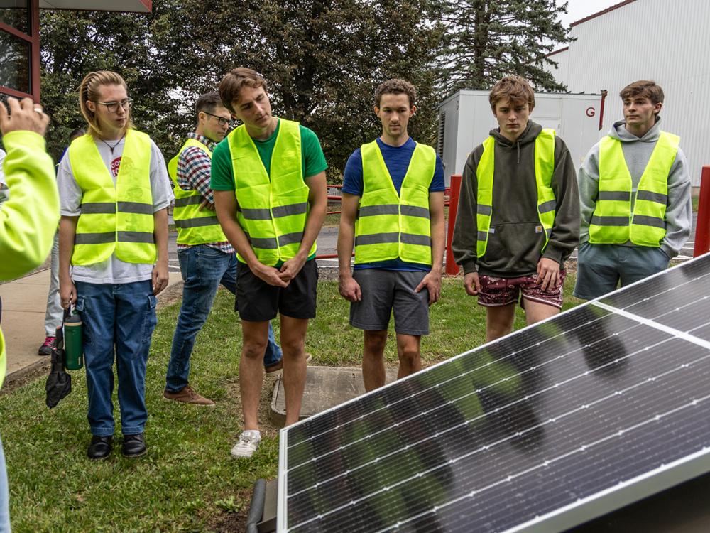 A group of young people in high-visibility vests attentively observe a solar panel outdoors.