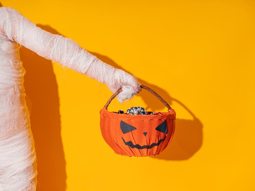 Mummy costume arm holding a pumpkin basket filled with candy against a bright orange background.