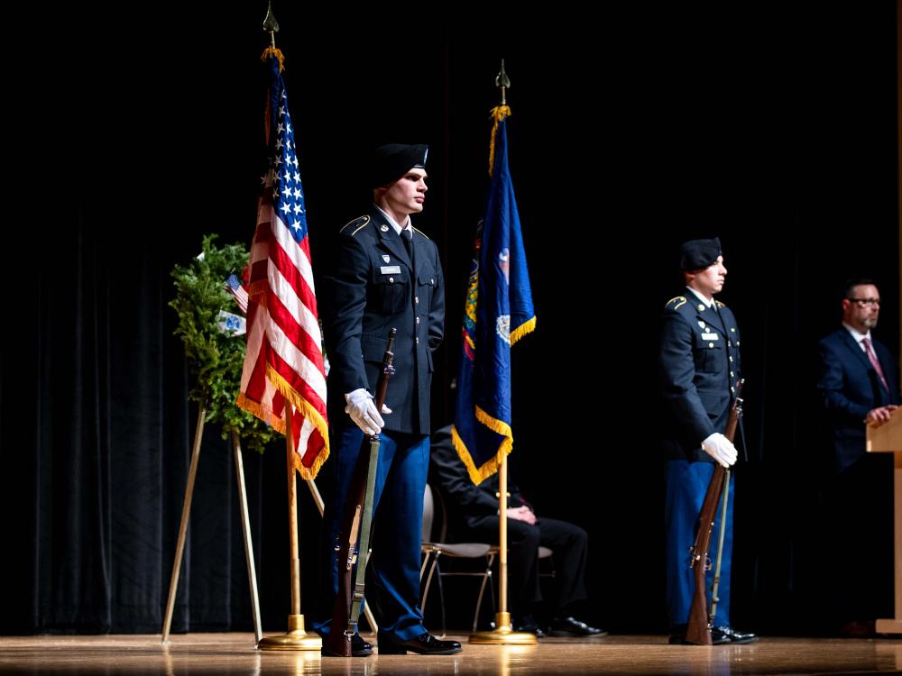 Two ROTC members in formal military uniforms stand at attention on stage