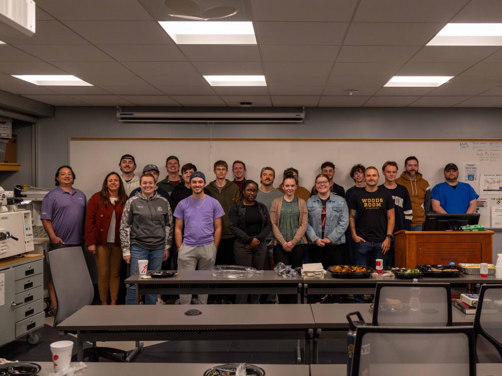 A group of about 20 students and alumni from Penn State New Kensington’s biomedical engineering technology program stand together at the front of a classroom during an alumni mentoring event. They are posed in a single row in front of a whiteboard, with tables, chairs and leftover food trays visible in the foreground.