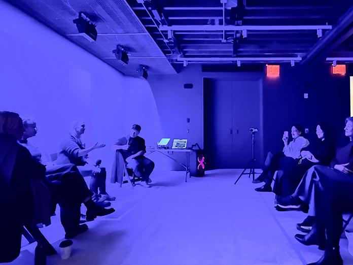 A blue-lit room with people sitting and watching a podcast being recorded