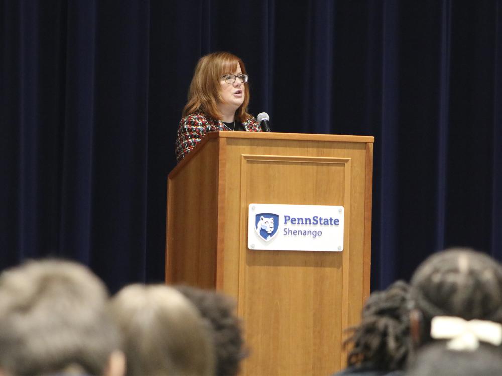 Mercer County Commissioner Ann Coleman speaks into a microphone behind a podium and middle school students sit in the audience.