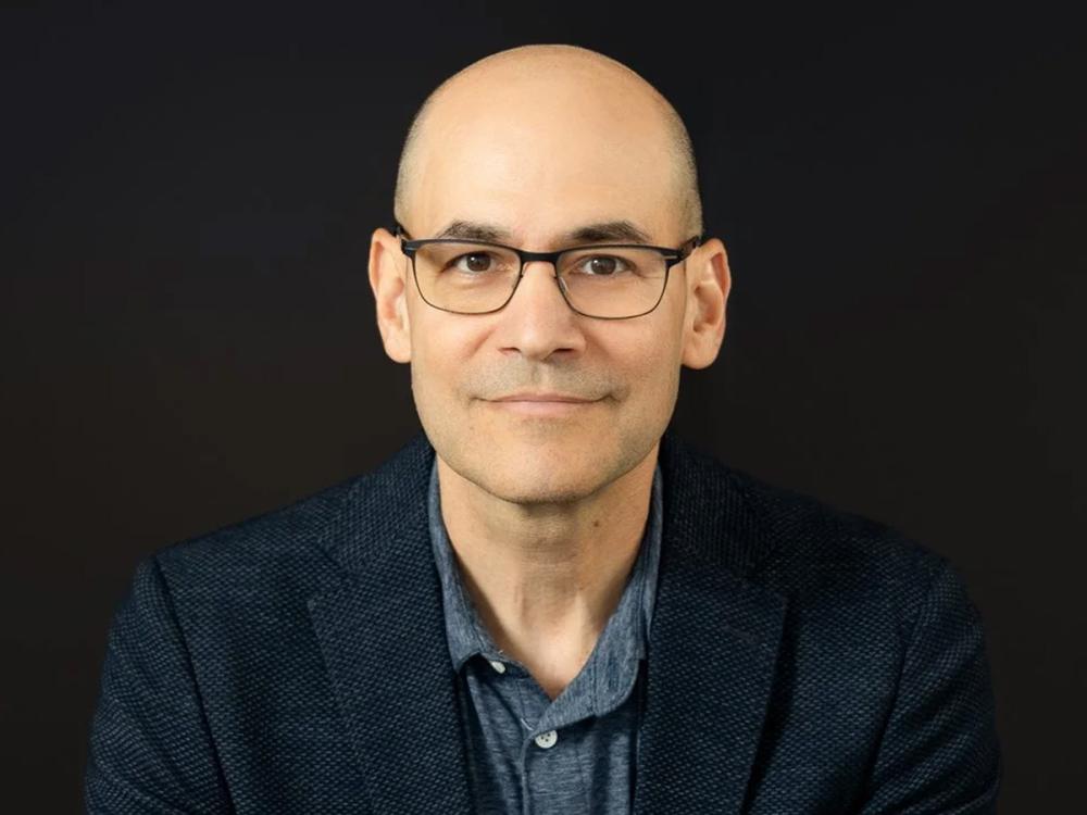 Benjamin DeAngelo is a bald man with glasses wearing a dark blazer and a blue shirt.