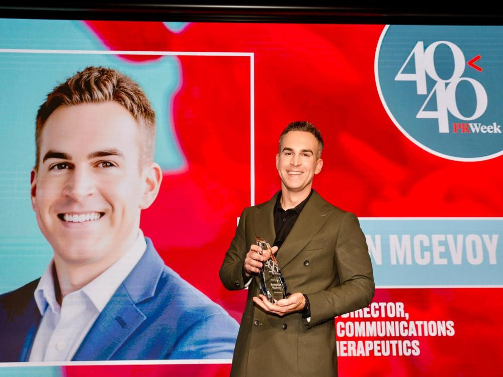 Man in a brown sports jacket holding a trophy in front of a red digital sign.