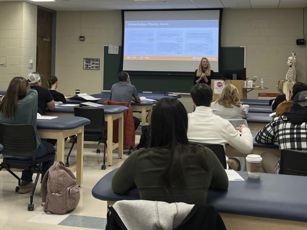 Roxanne Atterholt at the front of a classroom speaking to a group of people sitting at desks with a presentation projected behind her.