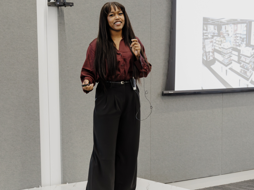 A student stands on a stage presenting with a microphone in hand and a screen on wall behind her