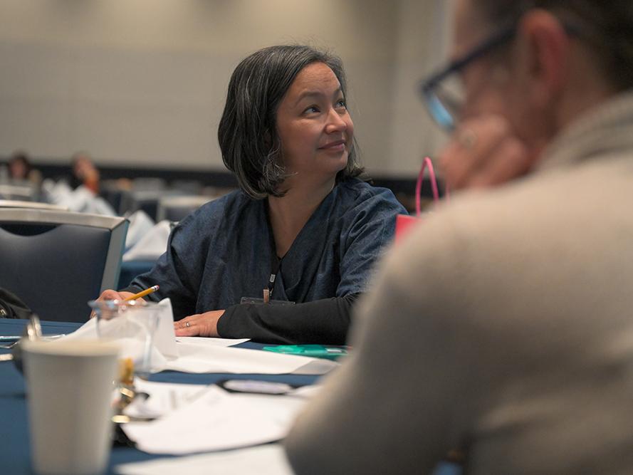 A woman with dark hair sits at a table and looks to her left.