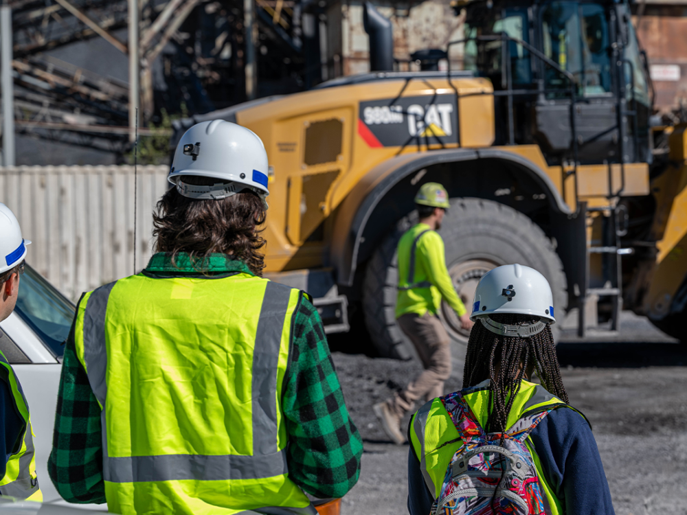 Students in safety gear at a construction site with a large yellow vehicle in the background.