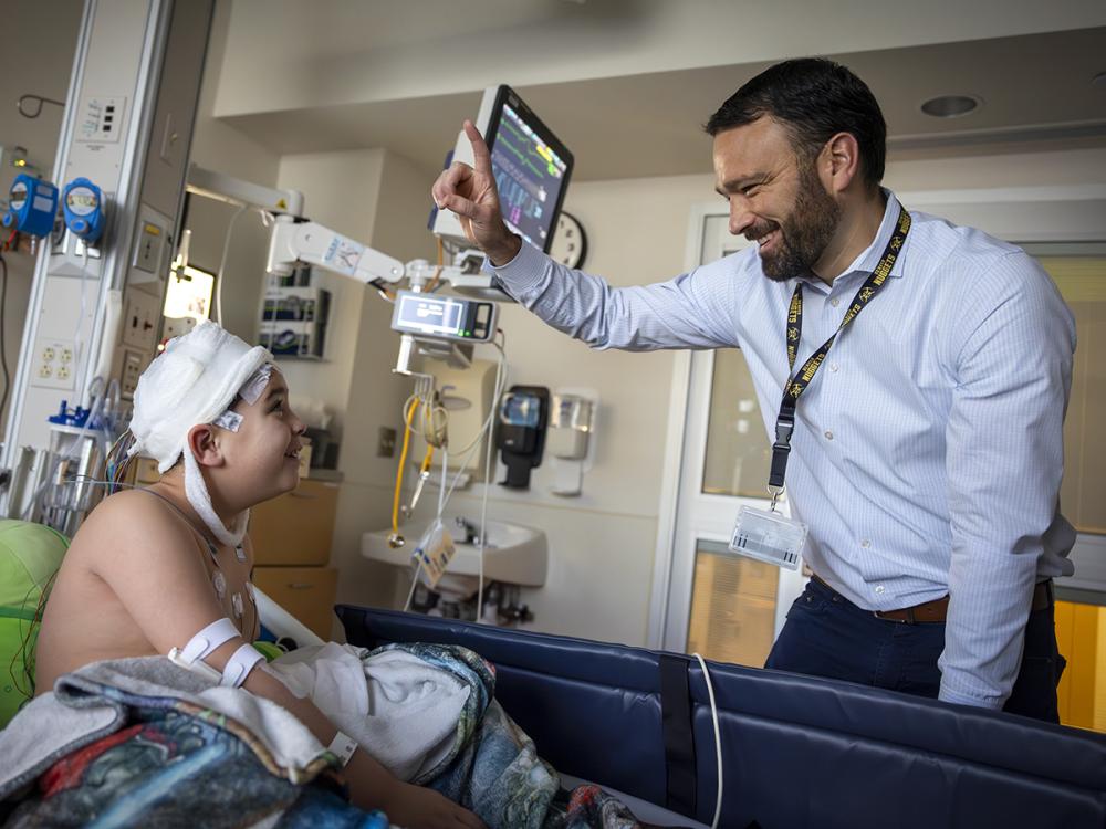 Brent O'Neill, right, wearing a shirt and tie, raises a finger and smiles at a boy in a hospital bed. The boy is looking up at him. The boy’s head is covered in bandages, he has an IV in his arm and has a blanket on him. Behind them are a monitor and medical equipment.