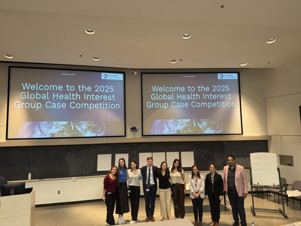 A large group of people stands in the center of a lecture hall below two projector screens.