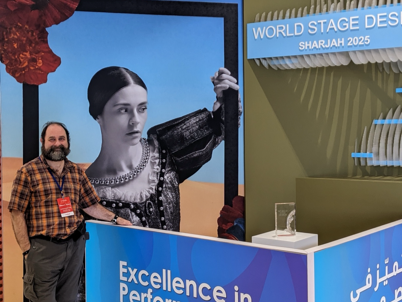 A bearded man wearing a plaid shirt and conference badge stands smiling beside a colorful exhibition booth. The booth displays signs reading ‘Excellence in Performance Design’ and ‘World Stage Design Sharjah 2025.’ Behind him is a large artistic image of a woman in historical-style clothing looking into a mirror frame.