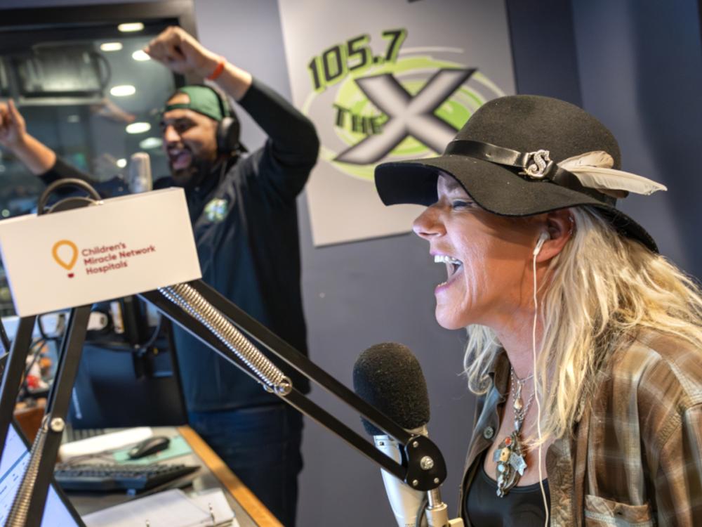 A woman wearing a black hat with feather accents laughs into a microphone while broadcasting in a radio studio. Behind her, a man wearing headphones and a backward cap raises his arms in celebration.