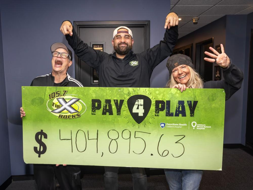 Three smiling radio hosts stand indoors holding a large green “105.7 The X Rocks Pay 4 Play” sign showing the fundraising total of $404,895.63.
