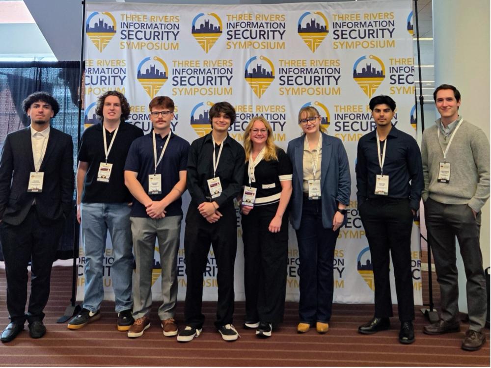 A group of Penn State Greater Allegheny faculty and students stand together in front of a banner reading “Three Rivers Information Security Symposium.” They are dressed in professional attire and wearing name badges, posing at the David L. Lawrence Convention Center in Pittsburgh.