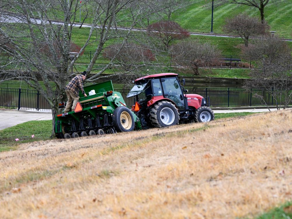 A large tractor pulls a seed spreader along an area of dead grass