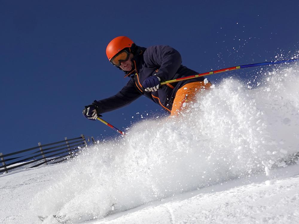 Male skier rides in the deep snow on a sunny day at a ski resort. He is wearing a helmet. A fence is on his left.