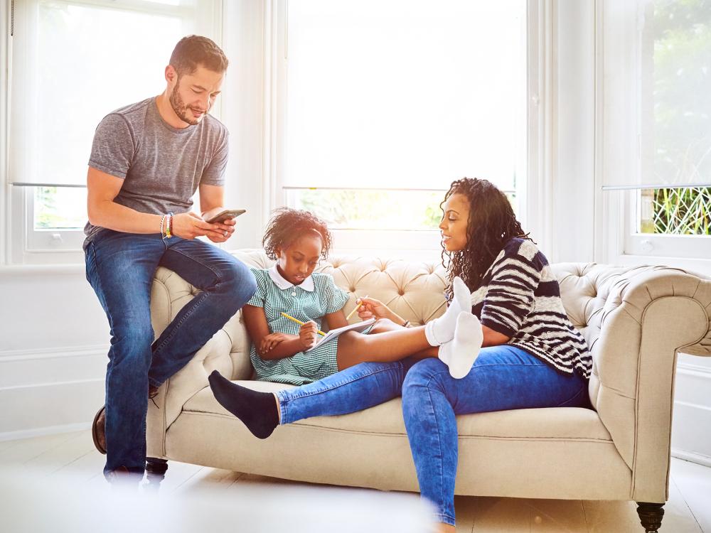 Family sitting on sofa in living room, with transgender male using mobile phone and female helping daughter in school homework