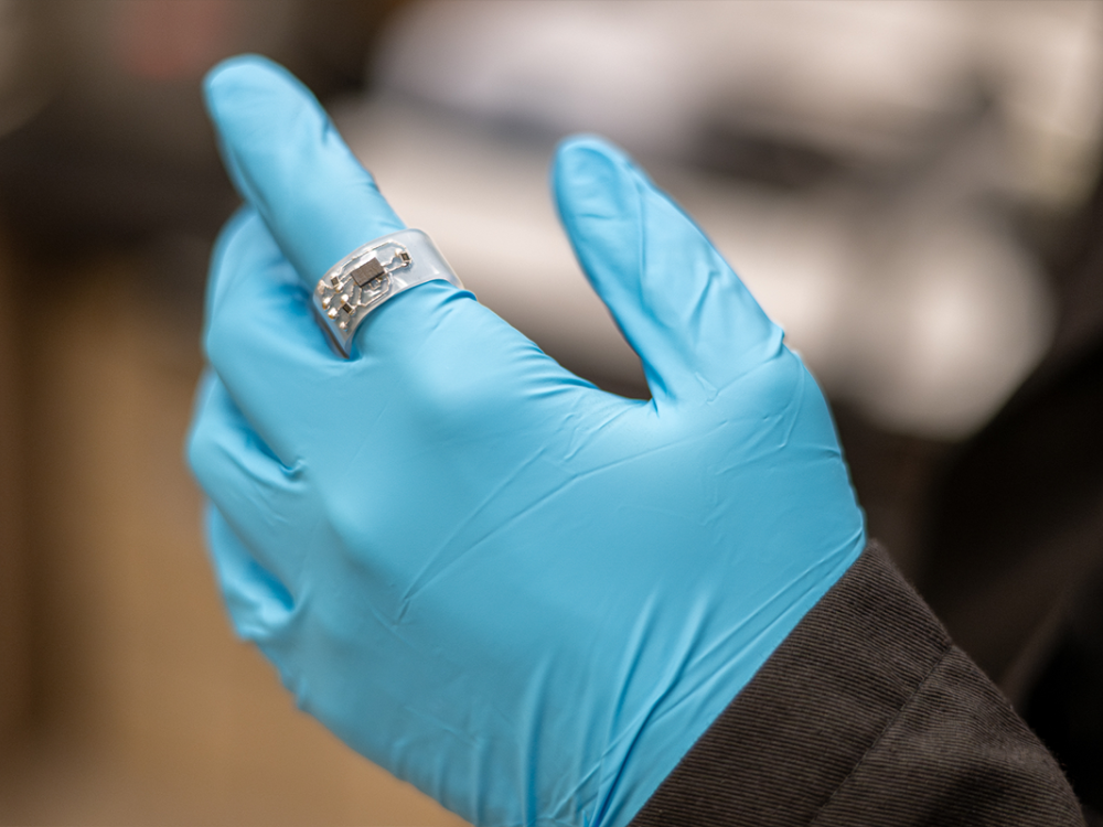 Clear ring with embedded circuit on a blue gloved hand in lab setting.
