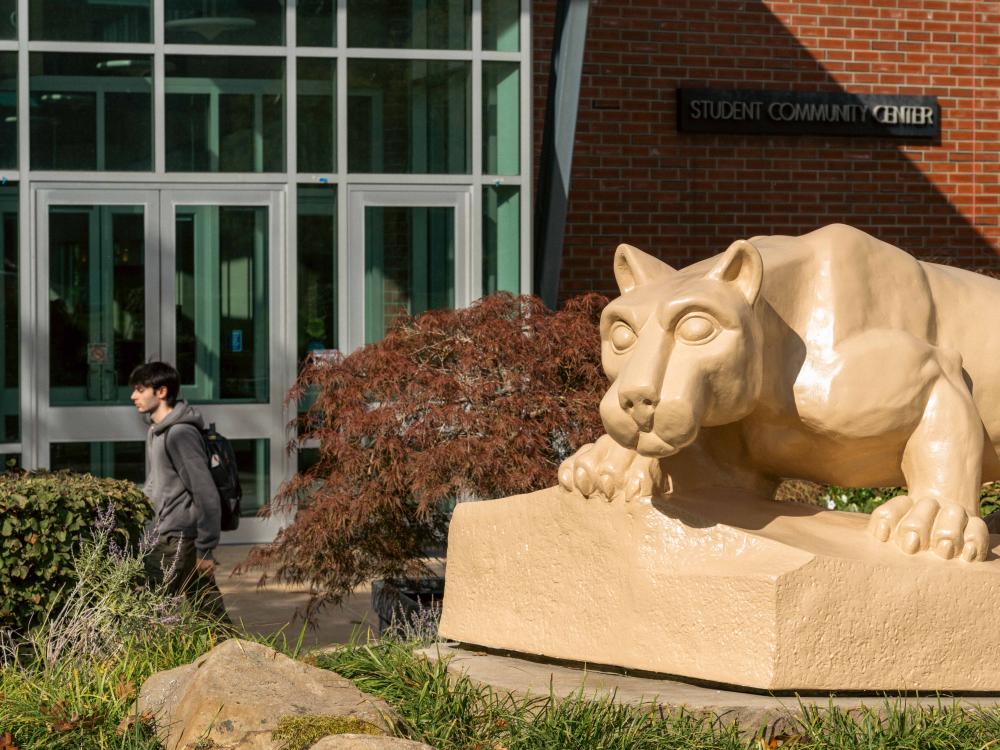 Nittany Lion statue crouches on a stone base in front of the Student Community Center at Penn State Greater Allegheny. A student with a backpack walks by on the left, and landscaped shrubs and plants fill the foreground.
