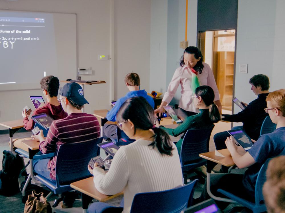 Kuei-Nuan Lin stands beside seated students in a classroom at Penn State Greater Allegheny, assisting them as they work on tablets during a mathematics lesson.