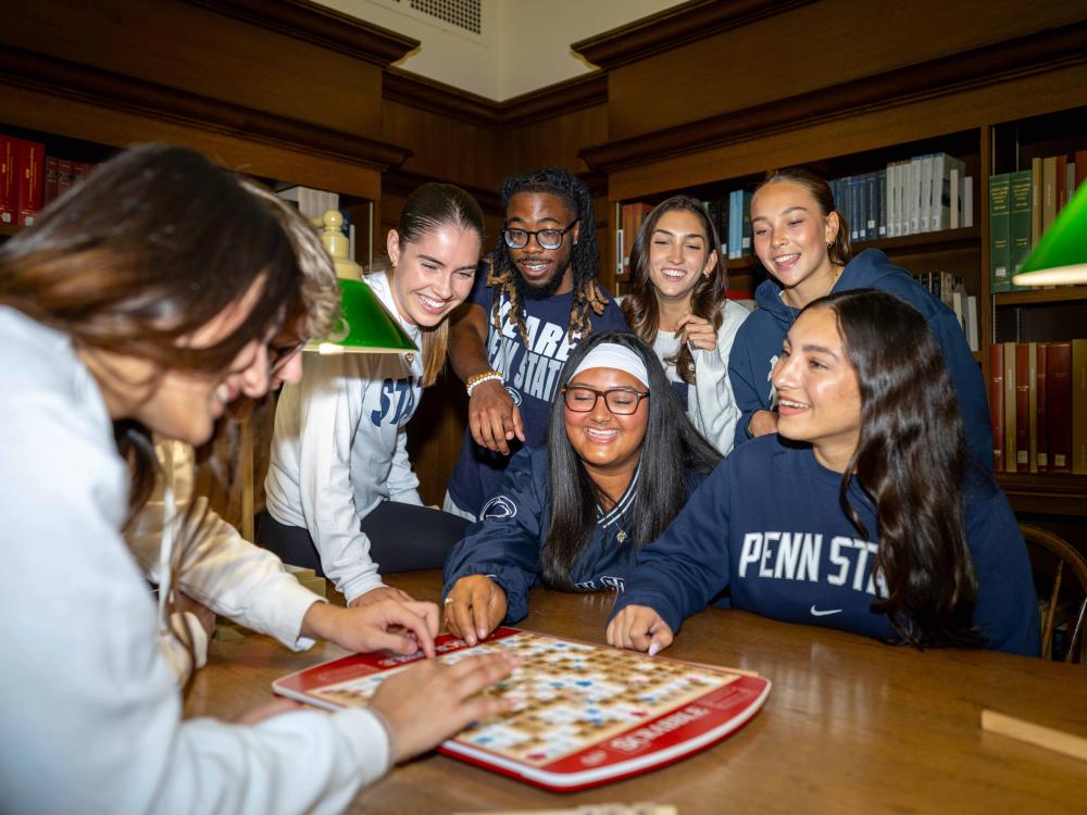 A group of students sits and stands around a wooden table in a library, leaning in toward a board game. They are smiling and interacting with the game pieces. Bookshelves line the background, and warm lighting from green-shaded lamps illuminates the scene.
