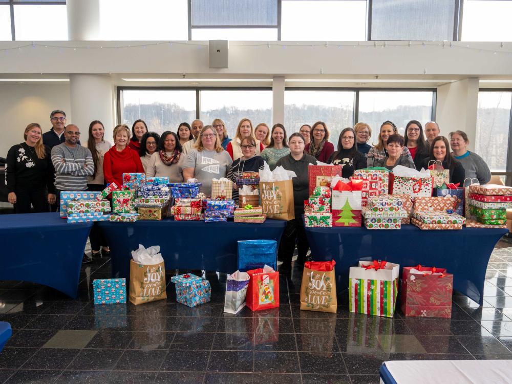 A group of more than 20 people stand behind tables laden with colorful, beautifully wrapped holiday gifts.