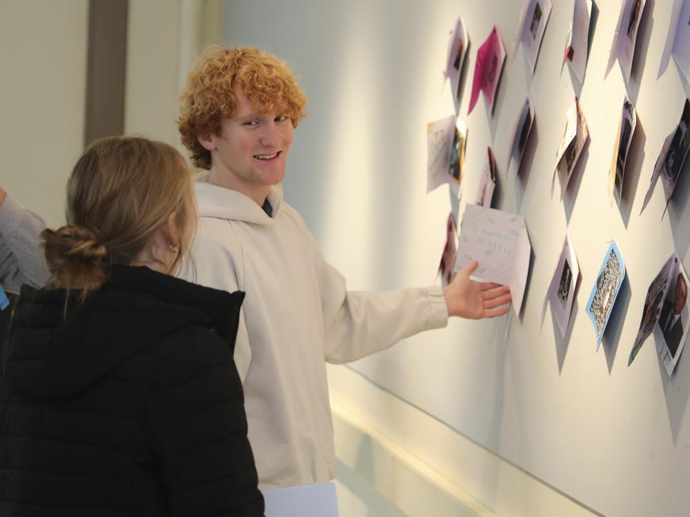 Two students look at each other and smile as one gestures towards art displays hanging on a wall.
