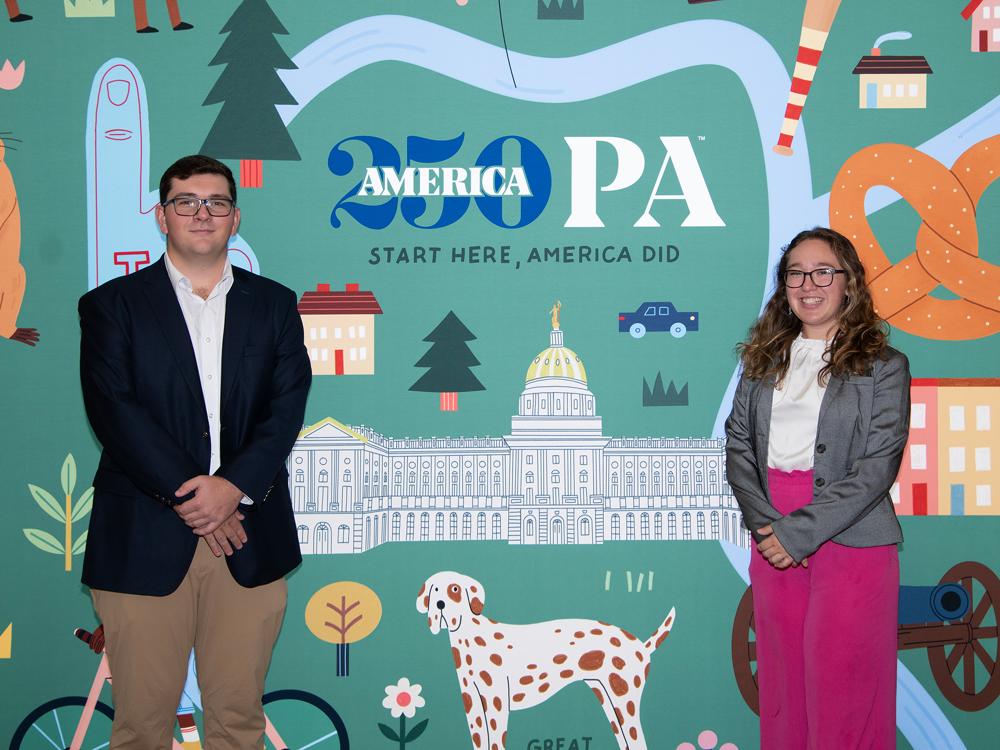 Penn State Altoona students Nick Riscica and Abigail Morgan stand smiling in front of a colorful America250PA backdrop featuring illustrations of Pennsylvania landmarks.