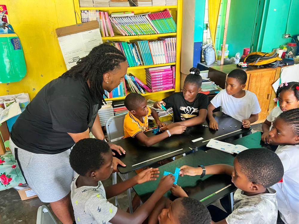 Instructor leans over a table helping a group of children work together on a hands-on activity in a colorful classroom.