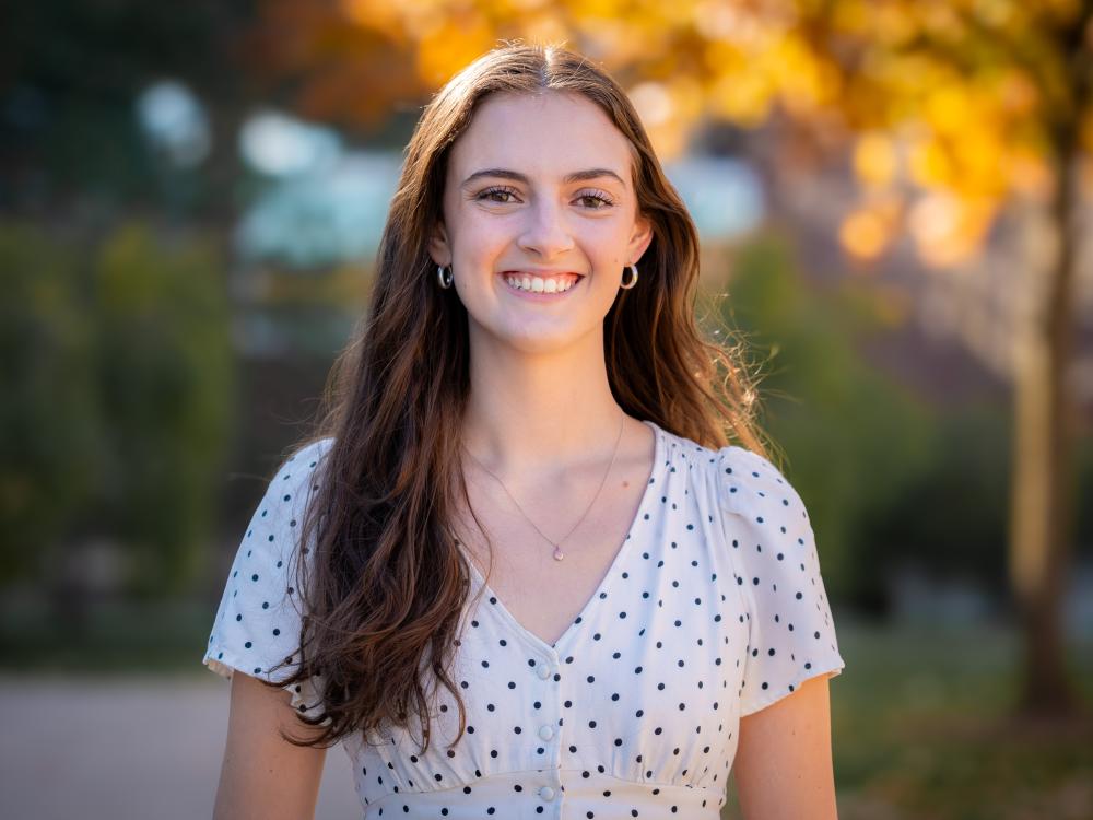 smiling person with long dark hair against backdrop of sunlit trees