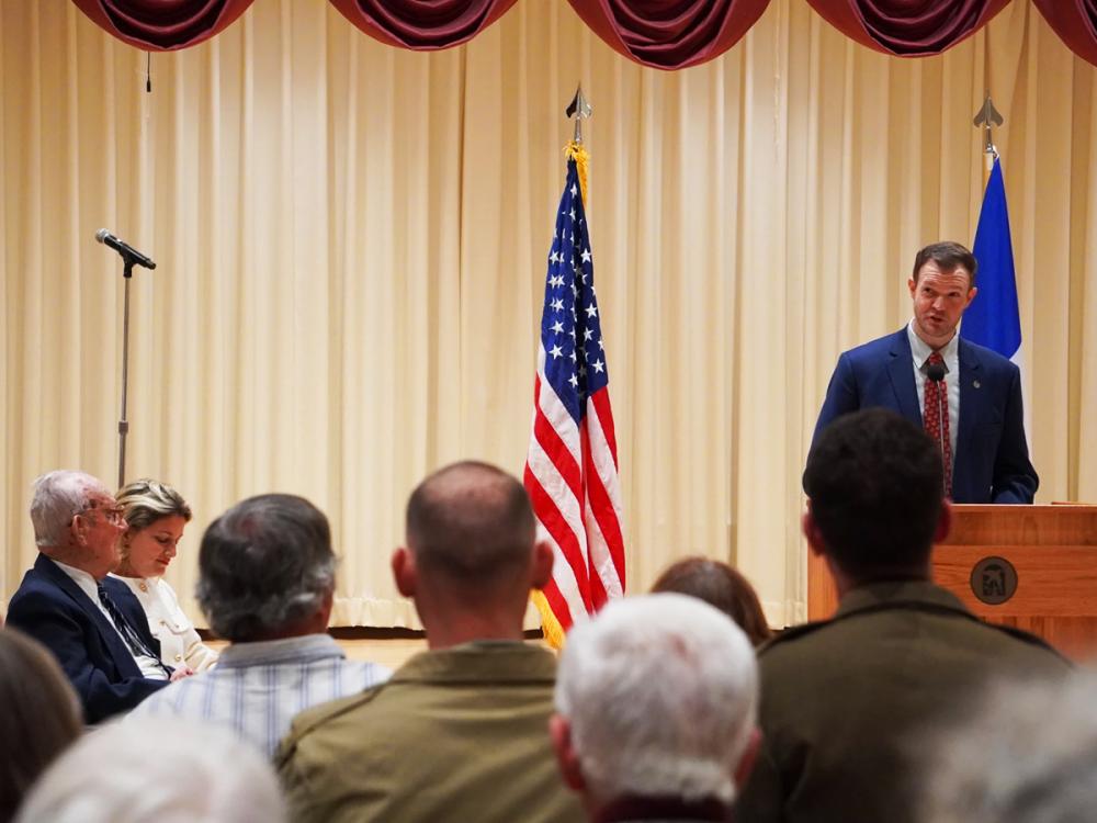 Jared Frederick at a podium addresses an audience in an auditorium, with U.S. flag and John Homan looking on.