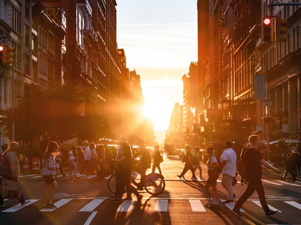People walk through a busy intersection in New York City on a summer day with sunset flare between the background buildings