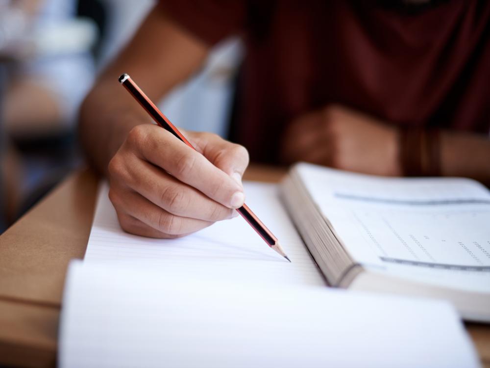 A close up of a person's hand holding a pencil, with an open textbook beside them