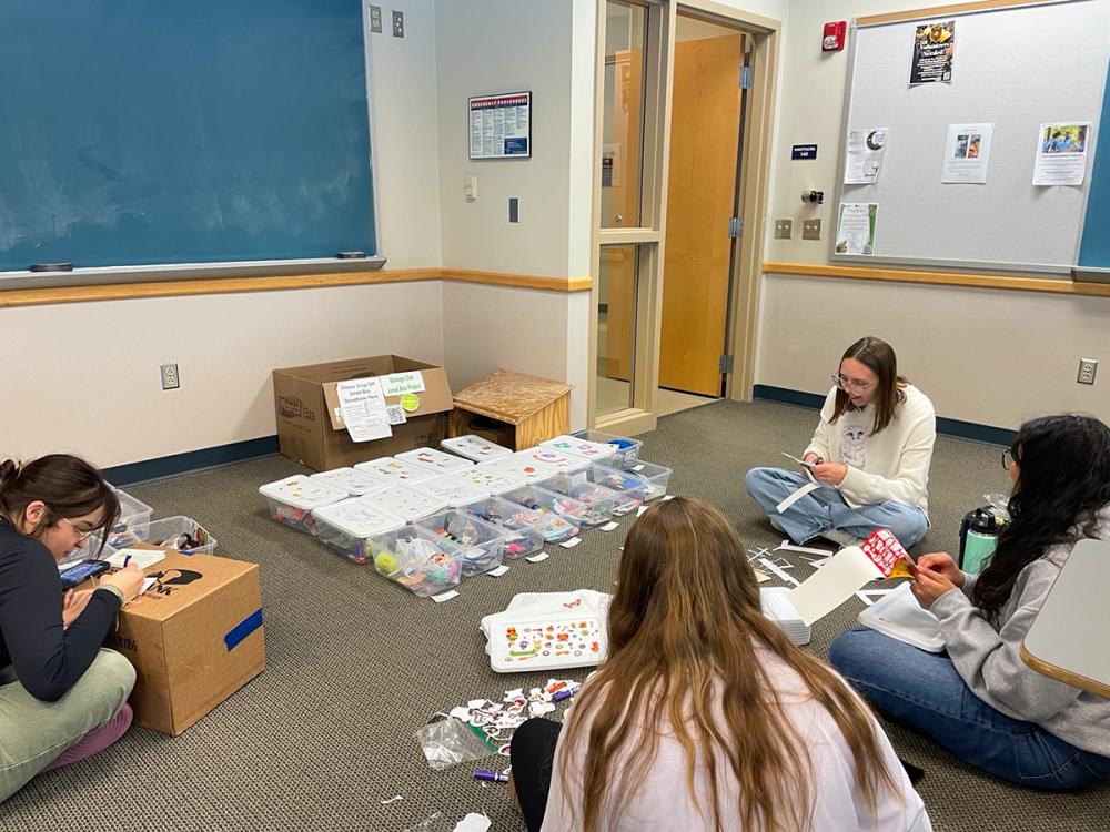 Students sit on a classroom floor organizing supplies and assembling kits from labeled bins during a group service project.
