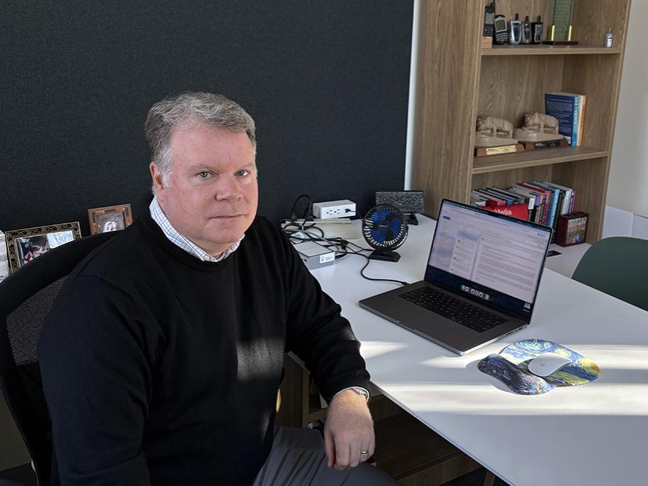 Man with short gray-ish hair wearing a black sweater sits at a computer desk.