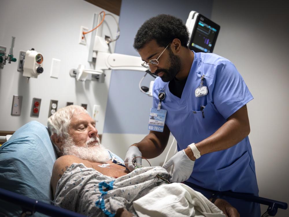 An ED technician tends to a patient laying in a bed. Various medical equipment is mounted to the wall in the background.