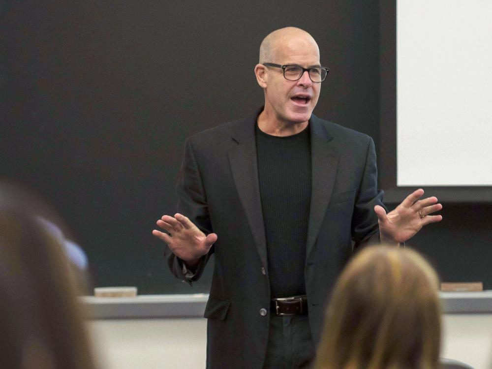 Bald man with glasses in a brown suit coat and black shirt gestures with his hands in front of a class